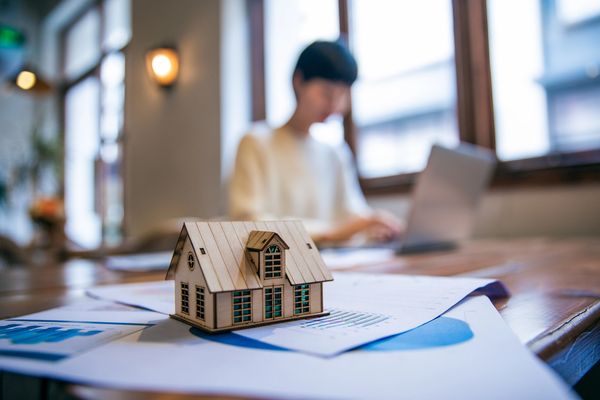 Miniature wooden house on financial documents with person working on laptop in background.