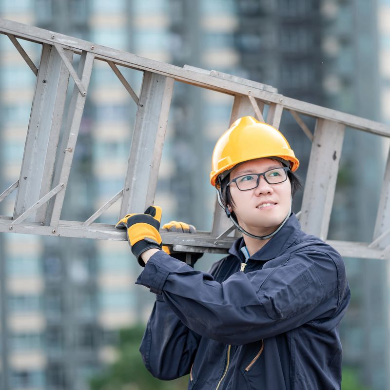 Asian maintenance worker man with protective suit and safety helmet carrying aluminium step ladder at construction site. Civil engineering, Architecture builder and building service concepts