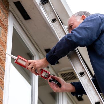 Man on ladder applying caulk around window frame.