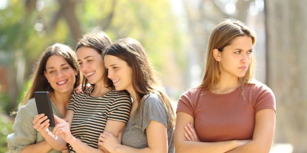 Four young women; three happily looking at a phone, one looks upset.