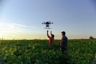 Two men operate a drone over a green crop field during sunset.