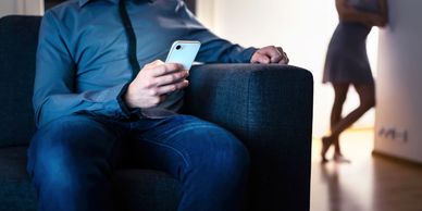 man looks at phone while woman stands in the background; representing infidelity problems