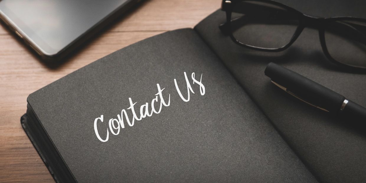 Notebook page with 'Contact Us' text, pen, glasses, and a smartphone on a wooden table.