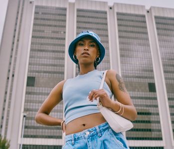 Confident young woman poses in blue crop top and bucket hat against tall building.