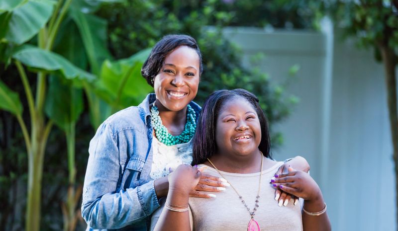 An African-American woman in her 30s standing with her teenage daughter, 16 years old, who has down syndrome. They are outdoors, in the back yard of their home, smiling at the camera.