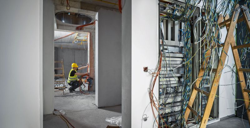 Electrician installing cables and corrugated pipes in wall at construction site with distribution board in foreground.