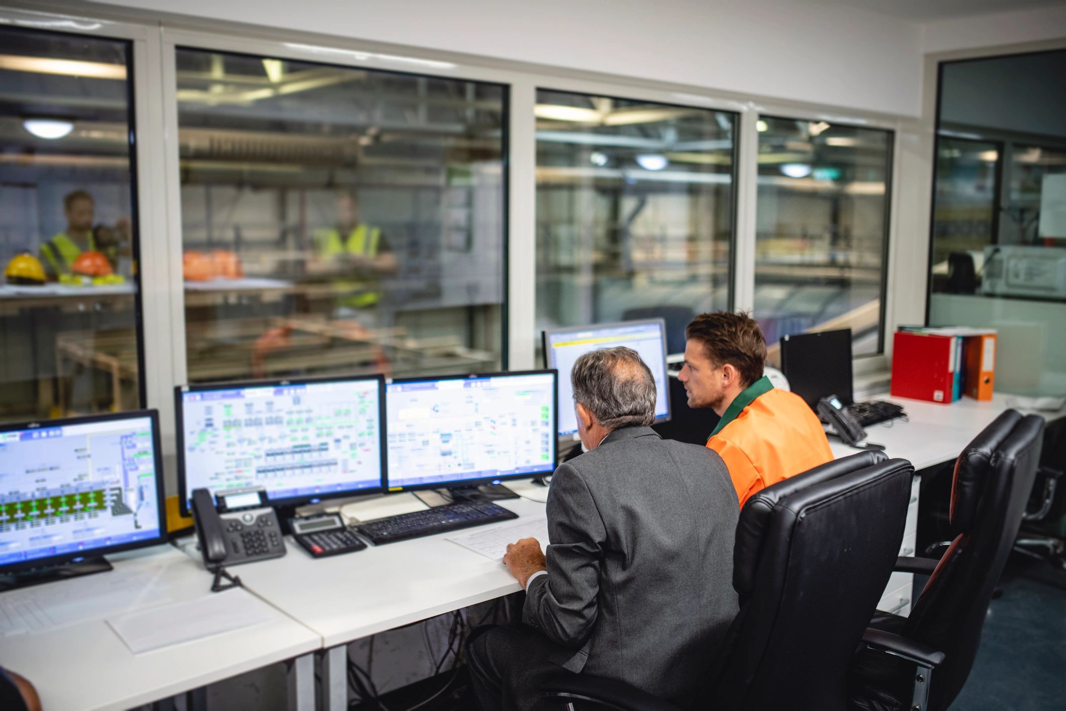 Rear view of male manager and worker in 70s and 30s sitting at desk with computers in control room of waste management facility.