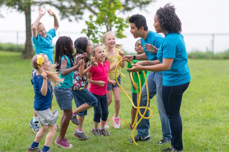 A group of children are jumping in excitement as they are getting ready with their camp counsellors to play a game of tug-of-war at summer camp.