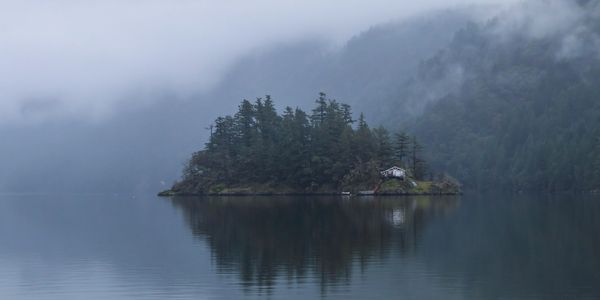 A small misty island with a cabin surrounded by calm water and forested mountains.