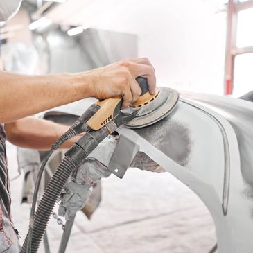 Worker sanding a car part with a power sander in a workshop.