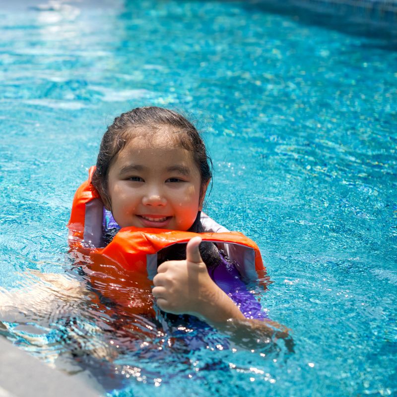 Asian child playing in the pool. Wearing orange life jacket, smiling with thumbs up.