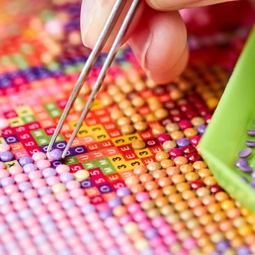 Close-up of a hand placing purple beads on a diamond painting canvas with tweezers.