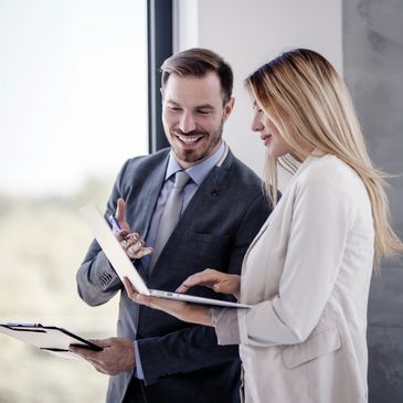 Two professionals discussing work with a laptop and clipboard indoors.
