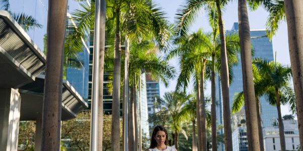 A woman walks through a palm tree-lined urban pathway with skyscrapers in the background.