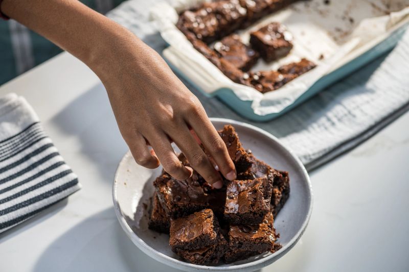 Cropped view of female hand stacking pieces of chocolate brownies into white bowl. Freshly baked squares of tasty dessert.
