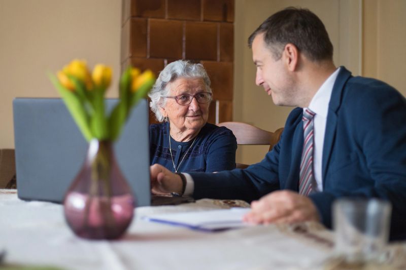 Real Estate Agent visiting happy elderly lady and showing her houses for sale on his laptop