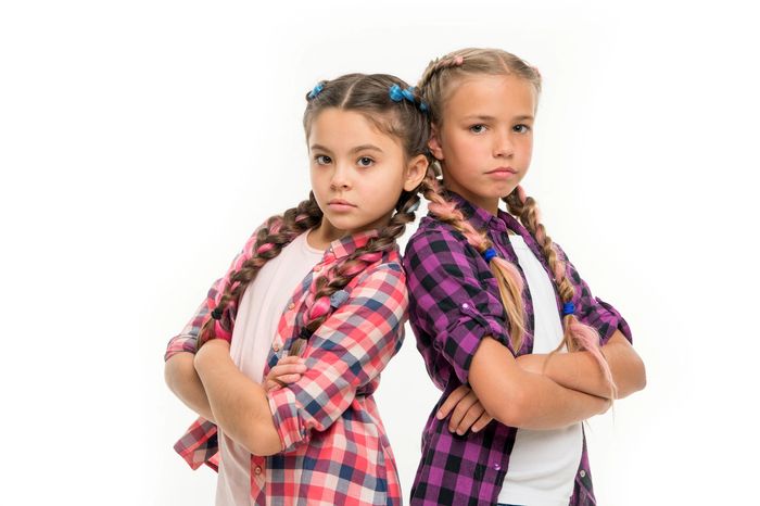 Two serious girls with braided hair and plaid shirts stand back to back with arms crossed.
