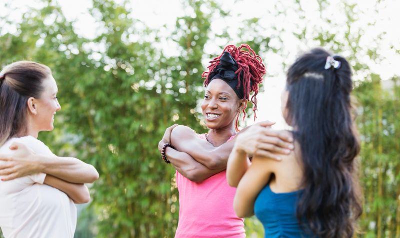 A multi-ethnic group of three women outdoors in a park, exercising. They are warming up, starting with some stretching exercises, standing in a circle facing on another. The focus is on the African-American woman, in her 30s with red locs.