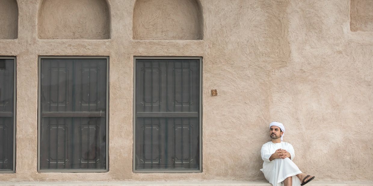 Man in traditional attire sitting against a textured beige wall with arched windows.