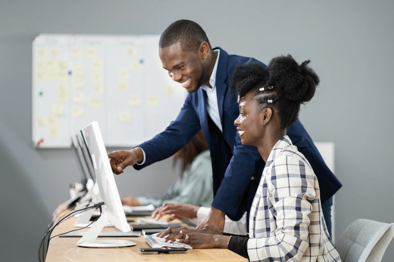 Two businesswomen are collaborating on a project, looking at a laptop and smiling, demonstrating teamwork and productivity in a modern office environment