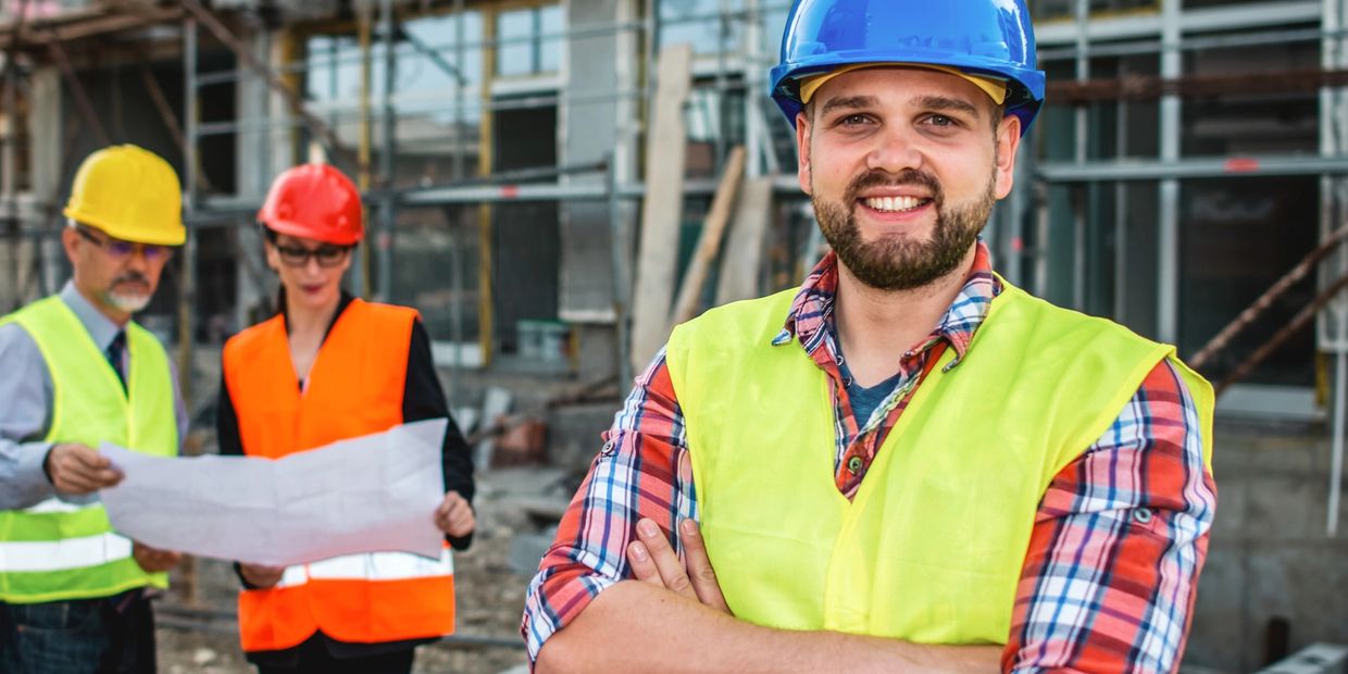 Smiling construction worker in blue helmet and vest with colleagues reviewing plans behind.