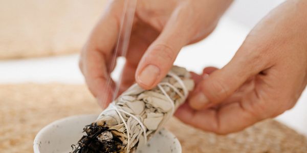 Hands holding a smoldering sage bundle over a ceramic dish on a woven surface.