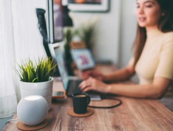 Woman working on laptop at wooden desk with smart speaker and coffee.