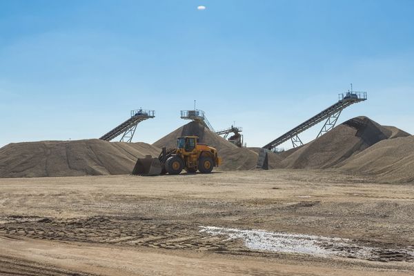Yellow loader in front of gravel piles and conveyor belts under a clear blue sky.