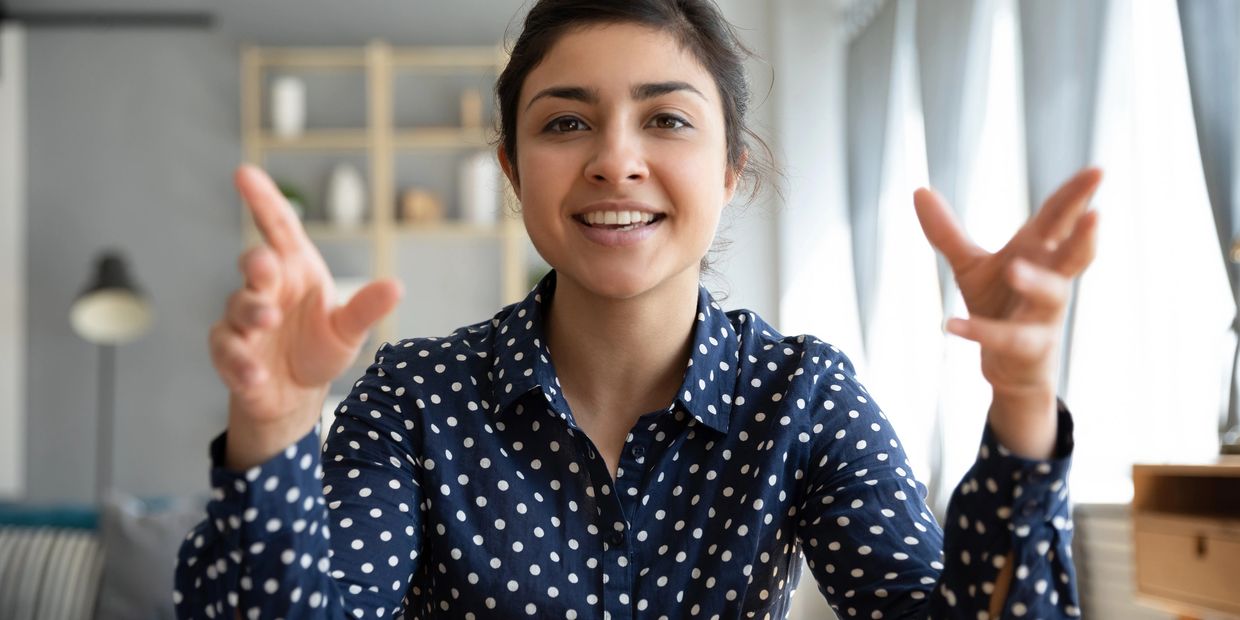Woman in polka dot shirt smiling and gesturing with hands indoors.
