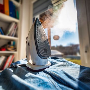 Close-up of a steaming iron on denim jeans near a sunny window.
