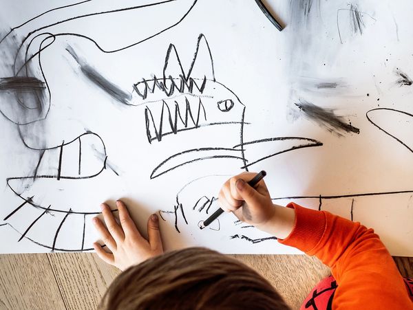 Child drawing a dinosaur-like creature with black crayon on white paper.