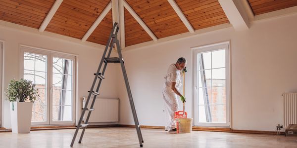 Man preparing to paint walls in a spacious room with wooden ceiling and large windows.