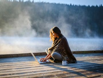 Woman wrapped in a blanket working on a laptop by a misty lake.