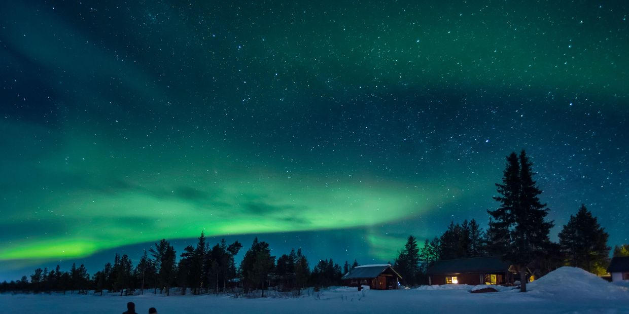 Northern lights glow over snowy landscape with cabins.