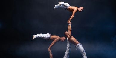 Four muscular acrobats perform a balanced human pyramid in white pants on a dark stage.