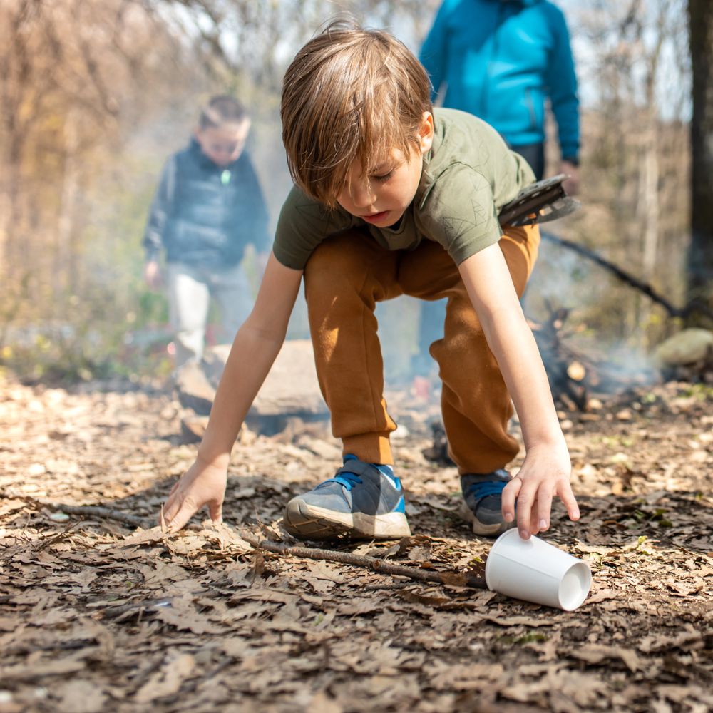Boy picking up a plastic cup in a forest with two others in the background.