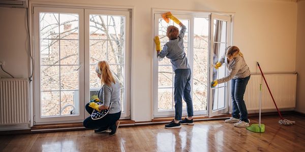 Three women cleaning large windows in a bright room with wooden floors.