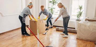 Three women cleaning and moving boxes in a bright, empty room.