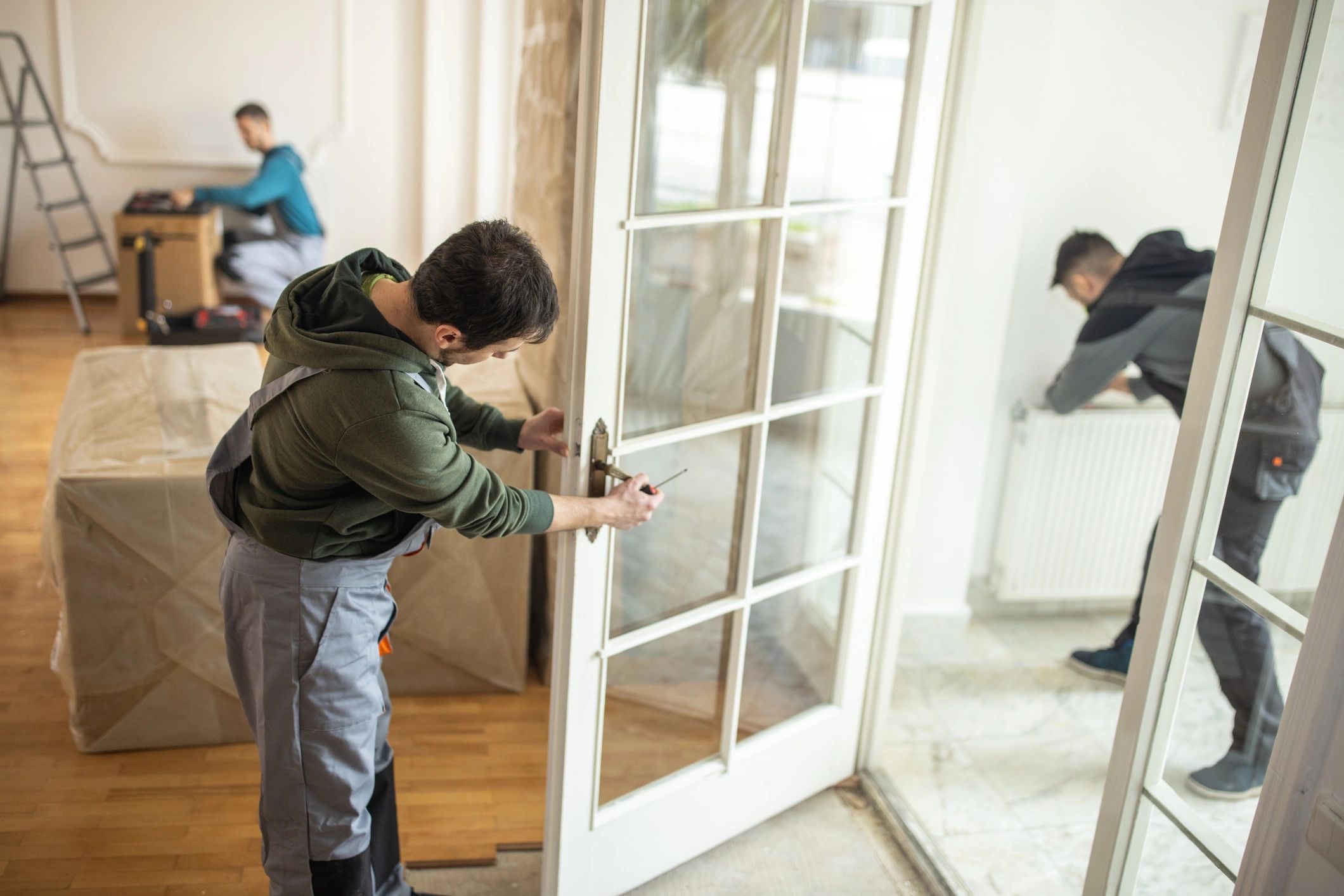 Three workers repairing a door and radiator in a bright room.