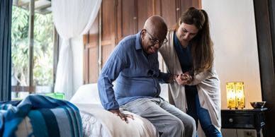 A woman helps an elderly man sit up on a bed in a cozy room.