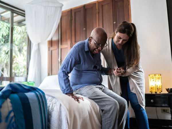 A woman helps an elderly man sit on a bed in a cozy room.