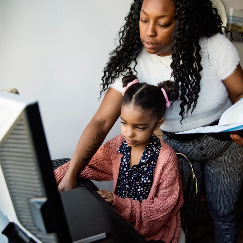 Cute mixed-race expressive little girl, doing homeschool sitting at the family desk computer. She is typing looking at the screen. Mother is helping behind her. Square indoors waist up shot with copy space.