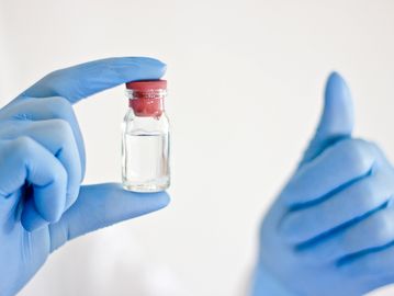 Gloved hand holding a small vial with a red cap, another hand giving a thumbs-up.