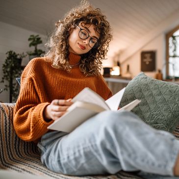 Young woman in orange sweater reading a book on a cozy couch.