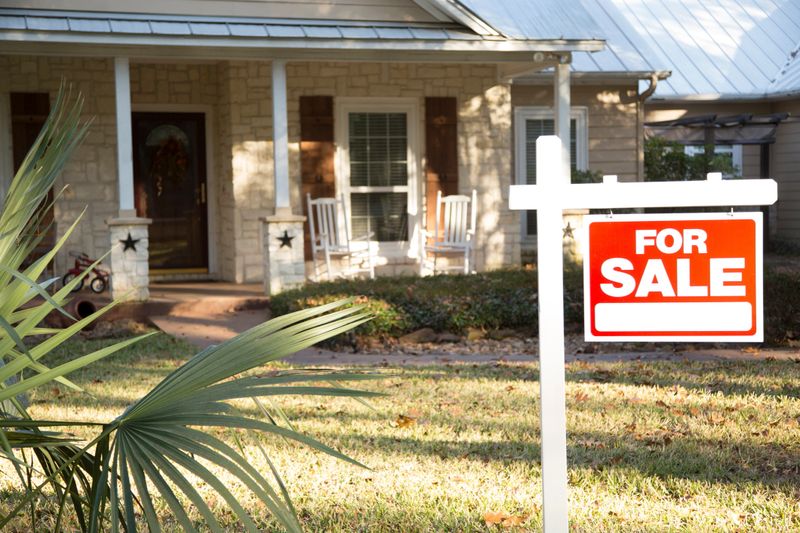 Home for sale with red and white real estate sign during the fall season.  Fall season with leaves on ground.  Front porch and windows in background.  Residential neighborhood.  Moving house, relocation concept.