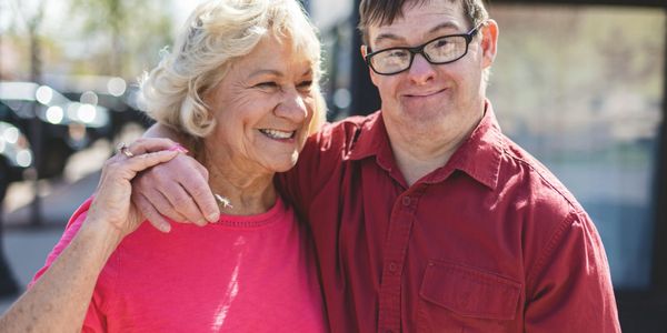 Smiling elderly woman and man with Down syndrome embracing outdoors.