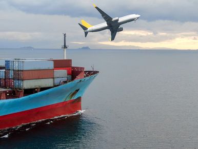 Cargo ship sailing with a commercial airplane flying overhead at dusk.