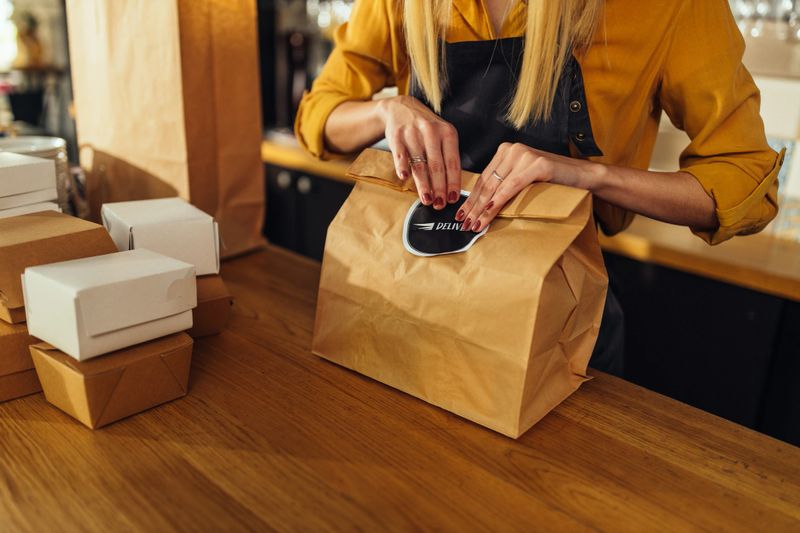 Close up of young woman packing food in paper bags and preparing them for delivery