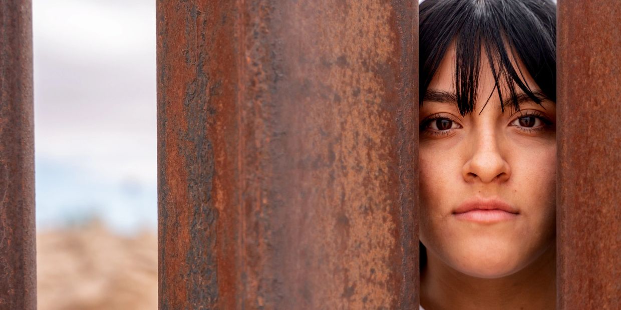 A woman looks through a rusted metal fence with a serious expression.