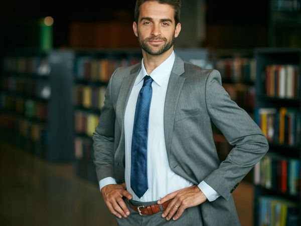 Confident man in a gray suit standing in a library with hands on hips.